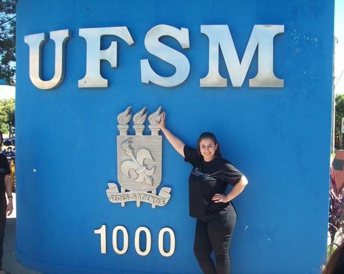 Foto horizontal da estudante vestindo preto em frente ao muro azul da UFSM com o brasão prateado