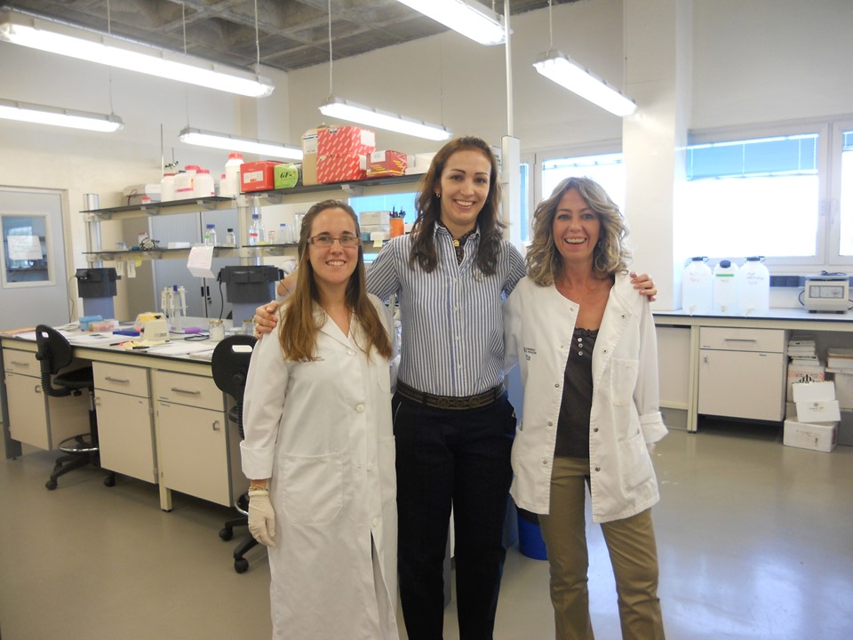 Foto horizontal com 3 mulheres em pé em um laboratório, duas vestem jaleco branco