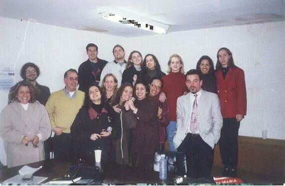 Foto horizontal com grupo de 14 pessoas, algumas sentadas outras em pé, numa sala branca
