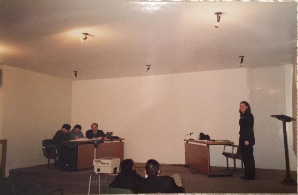 Foto horizontal, 3 pessoas sentadas em uma mesa e uma em pé apresentando trabalho