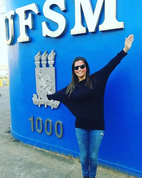 Foto vertical, mulher de braços abertos em frente ao muro azul na entrada da UFSM