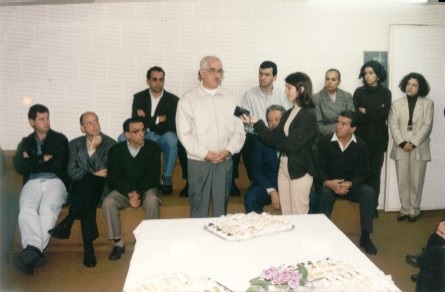 Foto horizontl com sala cheia de pessoas. Um homem e uma mulher mais à frente, o homem dando enteevista e a mulher gravando em um gravador portátil