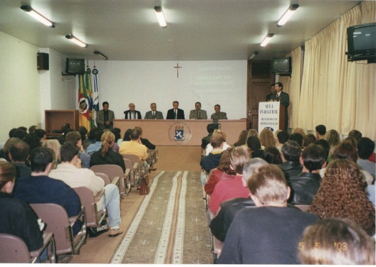 Foto horizontal, auditório cheio. Ao fundo palco e mesa com 6 pessoas sentadas e uma pessoa falando no púlpito