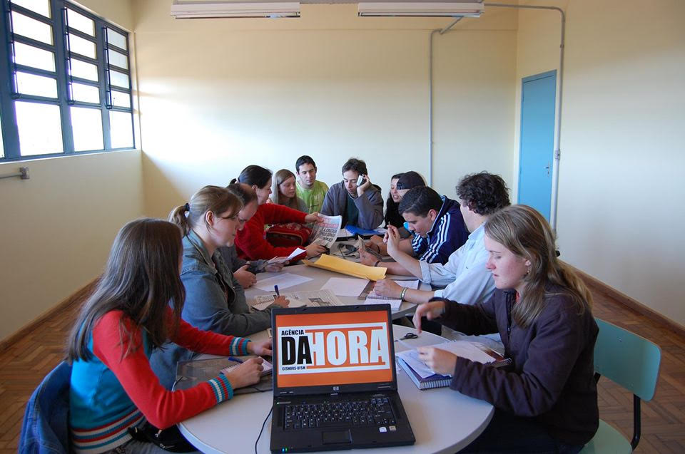 Turma reunida em torno de uma mesa oval, em primeiro plano notebook aberto escrito "Agência da Hora"