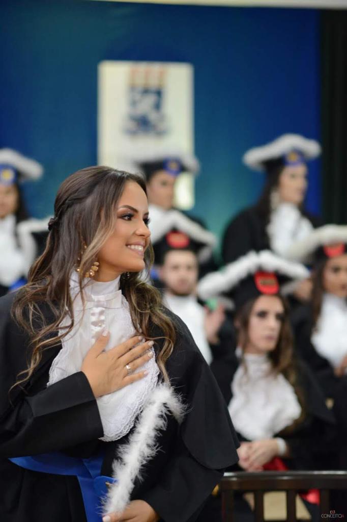 Estudante mulher em dia de formatura vestindo beca e segurando capelo, com turma de formandos ao fundoo
