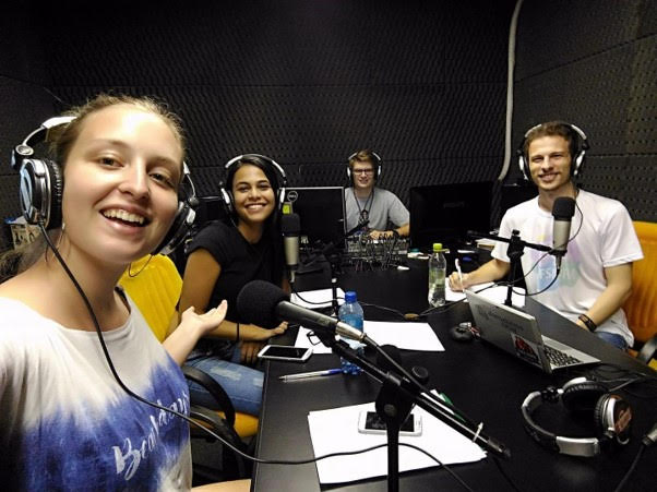 Foto horizontal em estúdio de rádio, alina em primeiro plano com mais 3 pessoas sentadas ao fundo