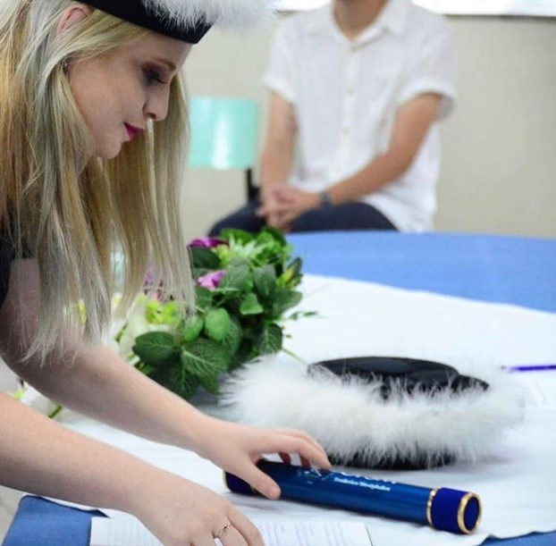 Foto quadrada, formanda usando capelo debruçada sobre mesa pegando canudo de formatura