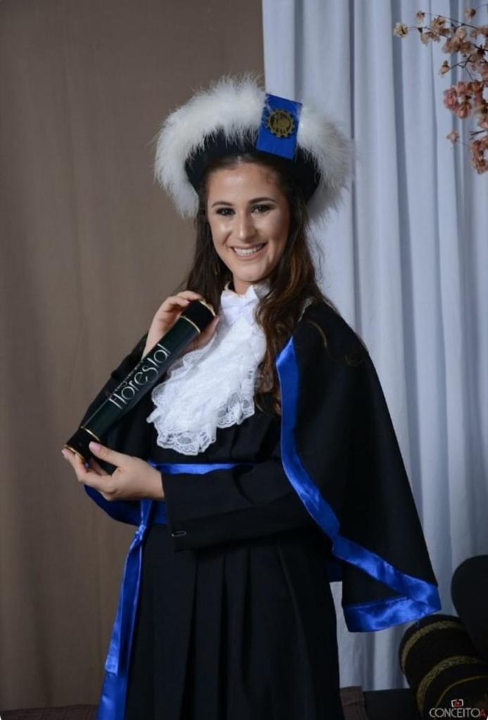 Foto vertical colorida de mulher vestindo beca de formatura e segurando canudo