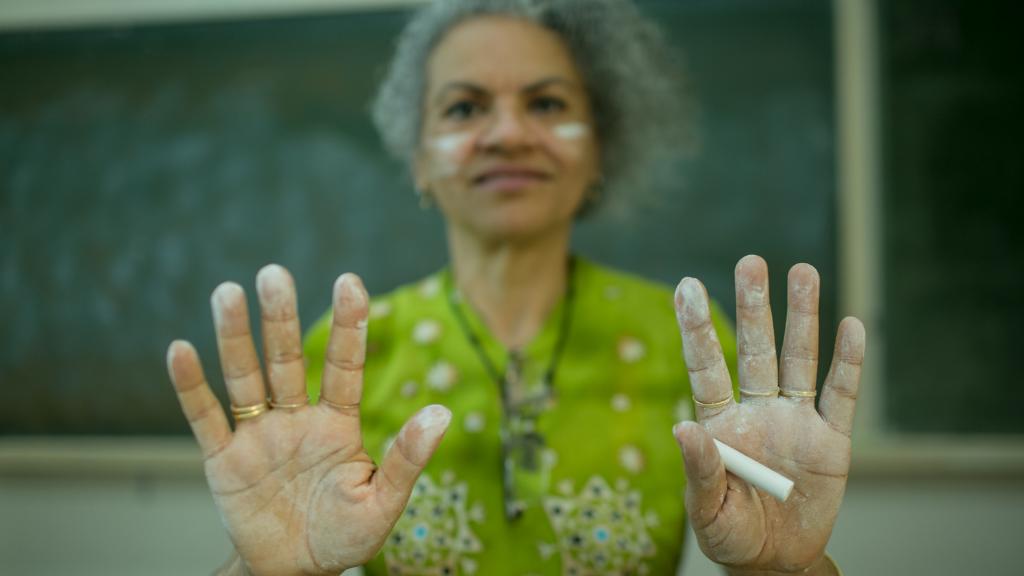 Fotografia colorida de uma mulher sorrindo com as duas mãos abertas e estendidas à frente do corpo, marcadas com giz