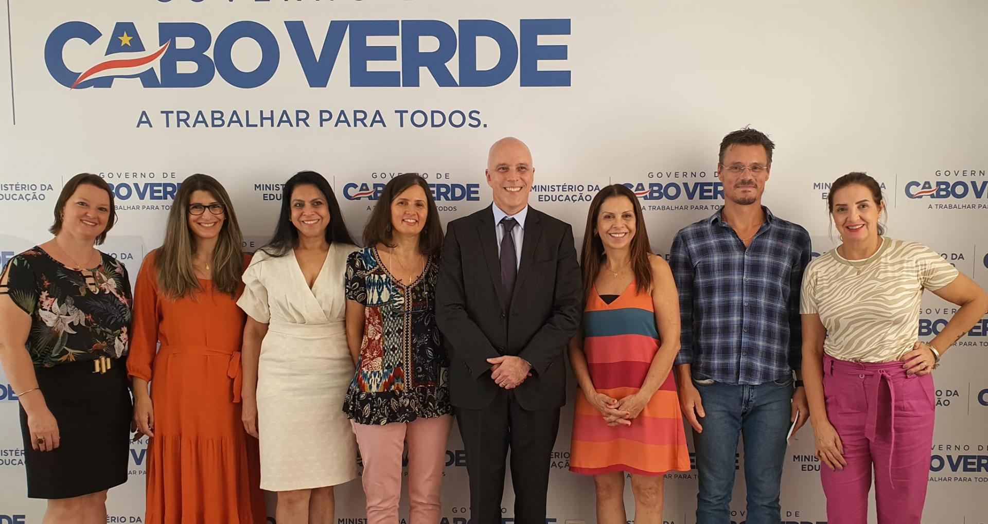 Descrição de imagem: na foto, aparecem oito pessoas de frente, em pé. Da esquerda para a direita, há quatro mulheres. A primeira tem cabelos curtos, veste uma saia preta com uma blusa estampada, com motivos florais, em tons brancos e cinza; a segunda tem cabelos louros escuros, na altura dos ombros, e está com vestido laranja; a terceira tem cabelos escuros e está de vestido branco; a quarta, de cabelos escuros, na altura dos ombros, veste uma calça rosa e blusa com estampas florais, na cor azul. Na sequência, está o embaixador, um homem calvo, de terno cinza escuro; a seu lado, há uma mulher de cabelos louros escuro, com um vestido de listras largas horizontais, nas cores laranja, bordô e azul. A seu lado, há um homem alto, de cabelos escuros vestindo uma camisa xadrez e calças jeans. A seu lado, há uma mulher de cabelos escuros, presos, com blusa bege e calça rosa escuro.