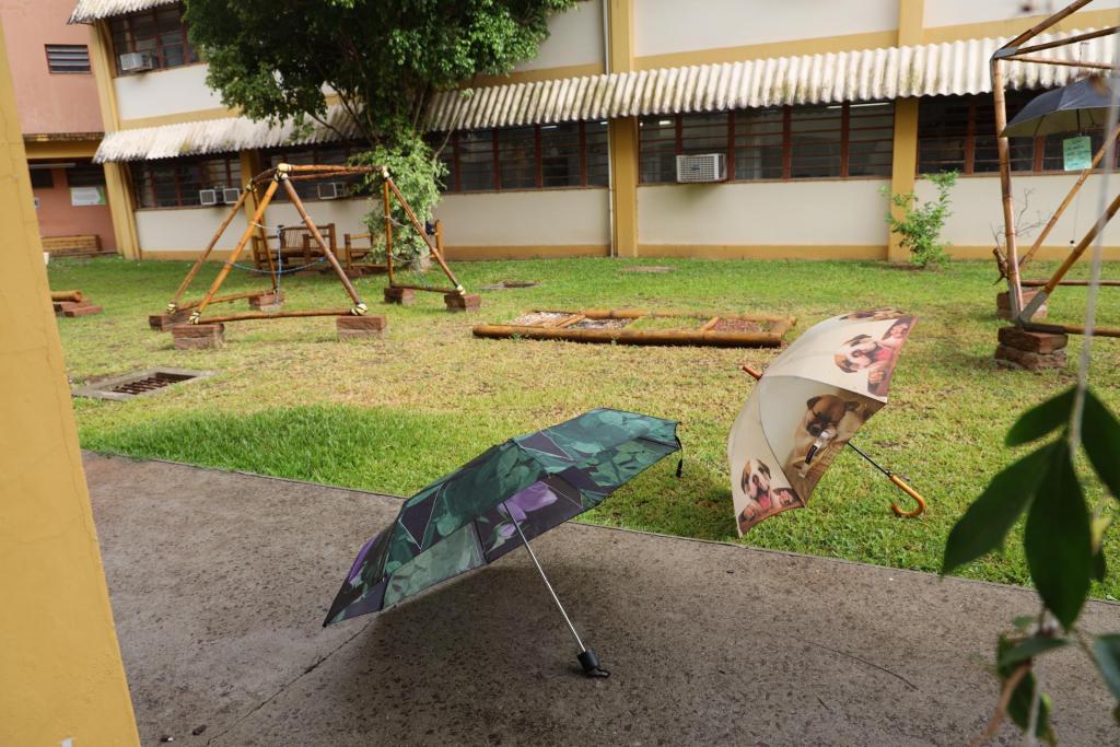 foto colorida horizontal mostra dois guarda-chuvas abertos no chão em um espaço de grama e calçada entre os prédios do CE