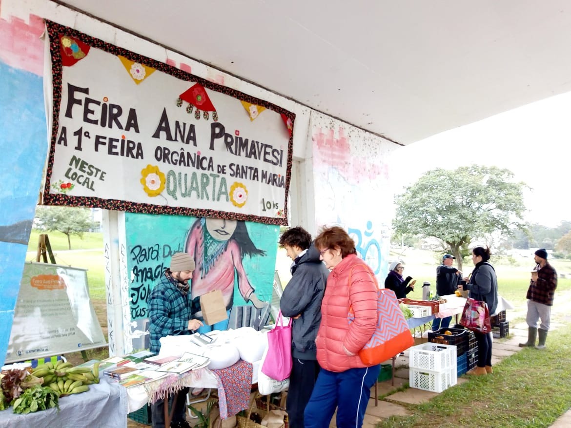 Foto horizontal. Cartaz com a frase “Feira Ana Primavesi, a 1ª feira orgânica de Santa Maria - quarta, neste local”, pendurado na cobertura da ponte seca da UFSM sobre uma pintura. Homem atrás de uma mesa com uma sacola de papel escolhendo algum item que está em sacolas brancas a sua frente. A frente da mesa um homem de casaco cinza escuro segurando uma sacola cor de rosa e ao seu lado uma mulher de jaqueta cor salmão segurando em seu ombros uma bolsa laranja, rosa e azul. 