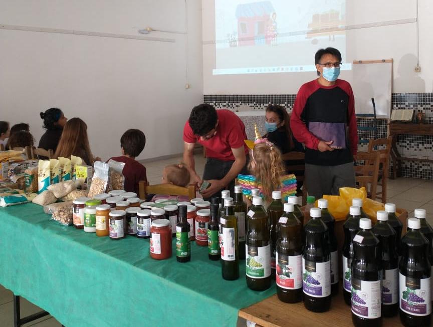 Foto horizontal. Mesa com uma toalha verde, composta por garrafas que contém suco de frutas, do lado direito há garrafas com suco de uva, abacaxi e morango. Do lado esquerdo há recipientes de vidro com doces e alguns pacotes de alimentos, atrás da mesa há várias pessoas, entre elas: um homem usando máscara, vestido com uma camisa com detalhes pretos, no centro a cor roxa e manga comprida na cor vermelha, a sua frente um criança com uma fantasia nas cores azul, amarelo e rosa. No fundo, uma mulher com roupa preta, usando máscara cobrindo o nariz a boca, olhando para um homem a sua frente, que está abaixado, vestido em uma camisa na cor vermelha e calça jeans, manipulando um item, na foto há 6 crianças, sendo que três aparecem ao fundo da foto olhando para um telão, não  é possível identificar suas vestimentas.
