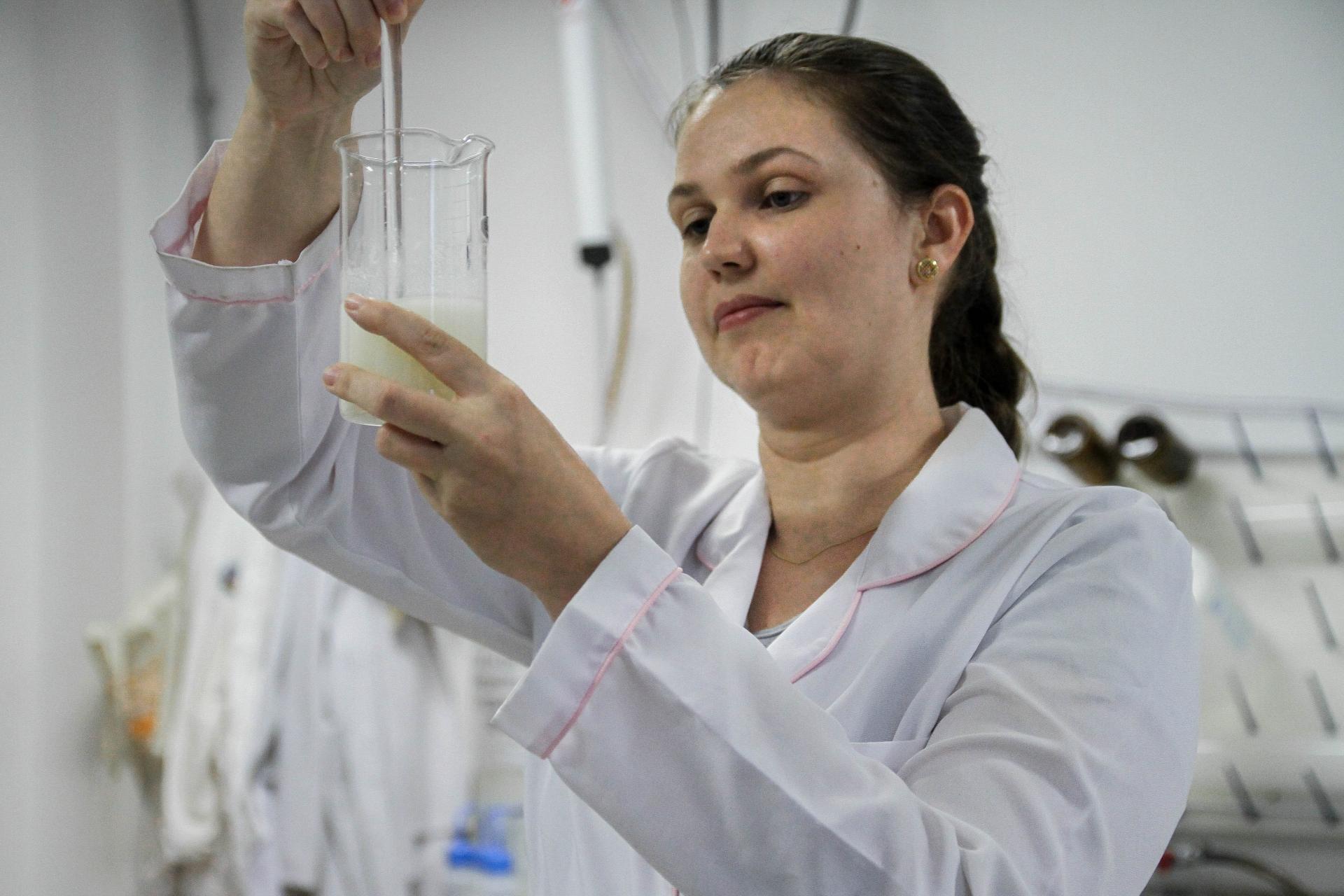 foto colorida horizontal mostra mulher, de jaleco branco, em um laboratório, com as duas mãos na altura do rosto manuseando equipamentos
