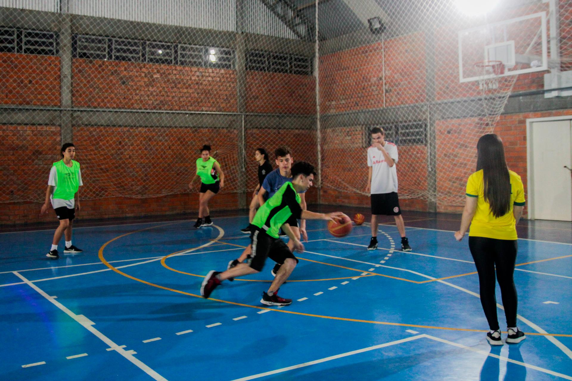 foto colorida horizontal com jovens jogando em uma quadra azul, em um ginásio de paredes de tijolo à vista. Alguns deles vestem colete verde limão