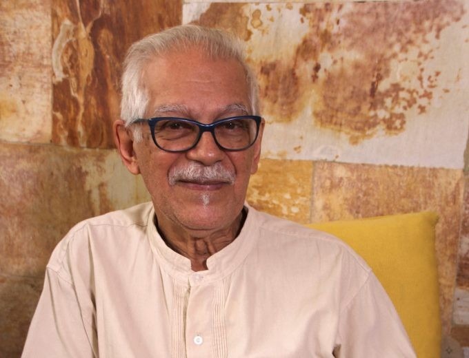 Foto colorida horizontal de homem idoso. Ele usa óculos, tem cabelo e bigode brancos, e usa uma blusa branca. Atrás dele uma parede em tons de amarelo e laranja. A imagem mostra do peito para cima.