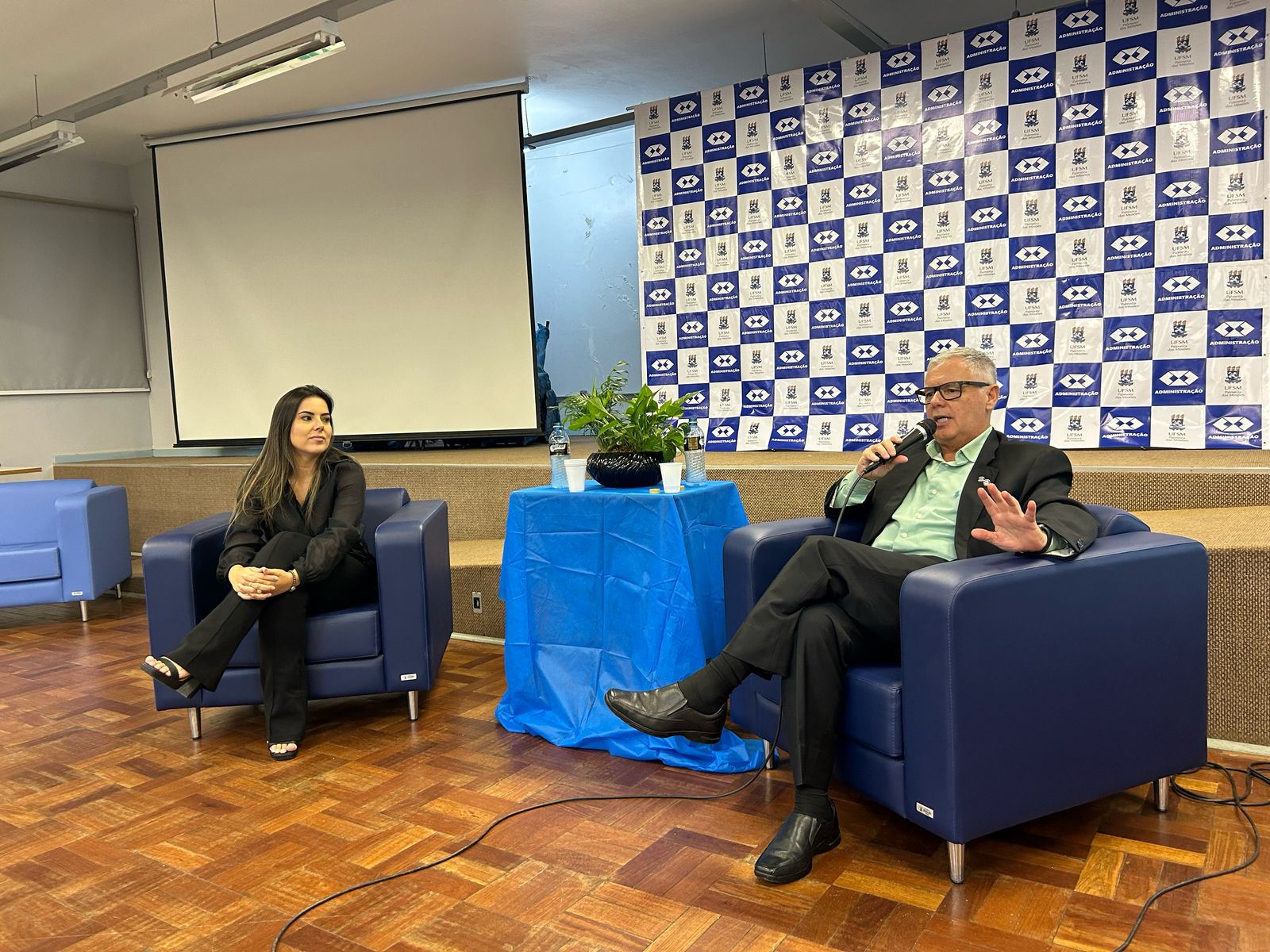 Foto colorida horizontal de duas pessoas sentadas em poltronas à frente de um painel com os símbolos da UFSM e do curso de Administração repetidos várias vezes. Na poltrada da esquerda, uma mulher jovem com as pernas cruzadas. Na poltrona da direita, um homem de meia idade, também com pernas cruzadas e microfone na mão direita.