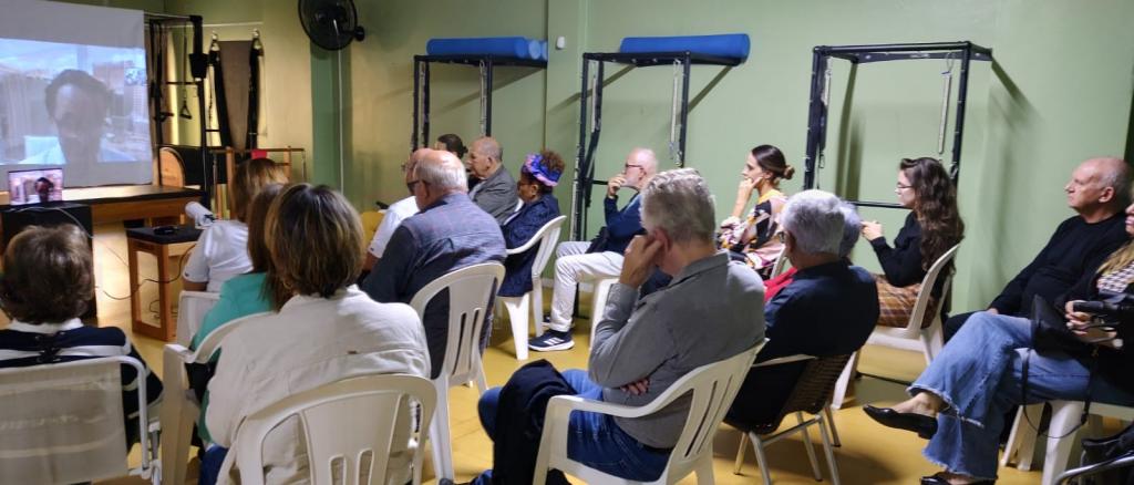 Integrantes do Movimento Parkinson estão sentados em cadeiras, em frente à uma tela que projeta o Dr. Alexandre Reis, durante a sua palestra no encontro especial de abril.