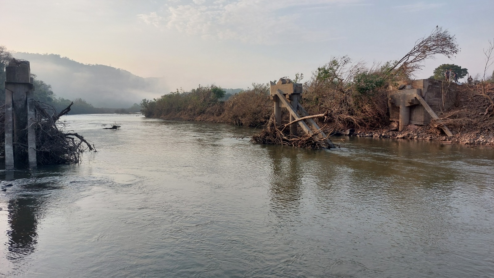 Foto colorida verticula de ponto destruída em Quevedos. Na imagem, apenas estão as cabeceiras da ponte e o rio. Não há, na foto, nenhum vestígio da ponto. Também há vários troncos presos nas cabeceiras.