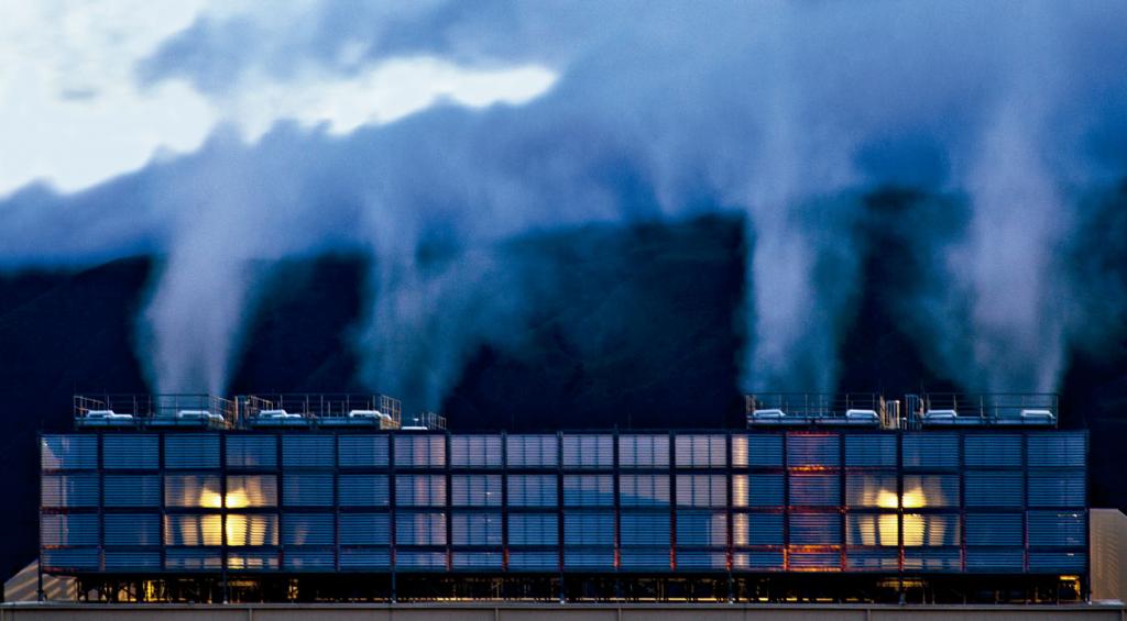 Foto colorida horizontal de um prédio sistema de resfriamento com grades metálicas largas, de onde saem várias colunas de vapor branco em direção ao céu. Ao fundo, colinas escuras.