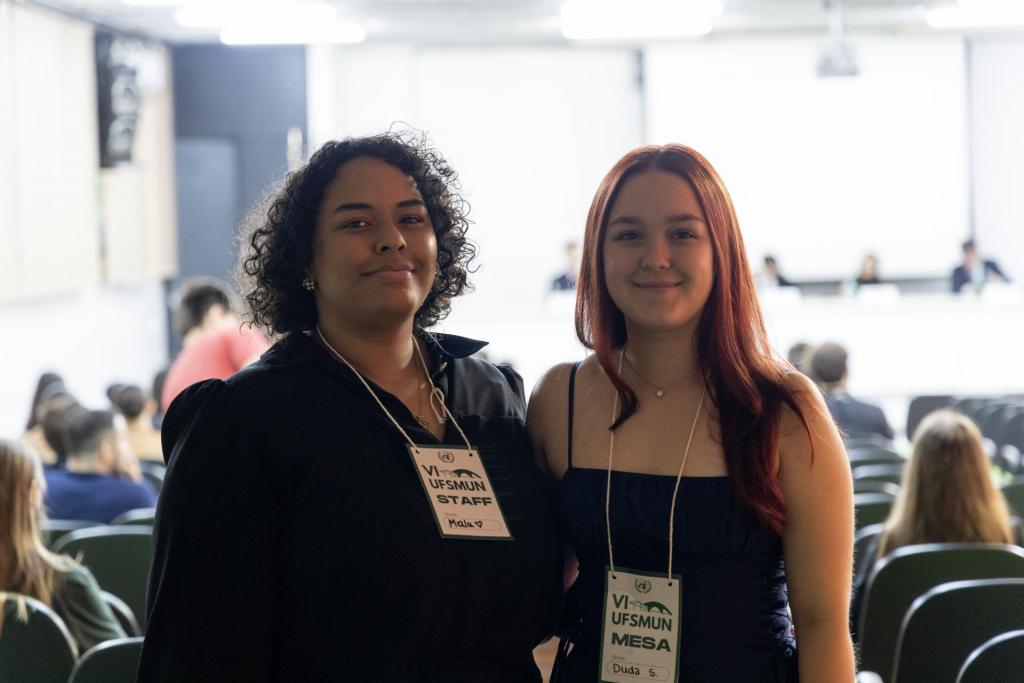 Foto colorida e horizontal. Em primeiro plano, duas pessoas estão em pé, sorrindo, em um auditório. À esquerda, uma pessoa de cabelos curtos e cacheados veste roupa preta; à direita, uma pessoa de cabelos longos e ruivos veste roupa azul-escura. Ambas usam crachás do evento VI UFSMUN. Ao fundo, há várias pessoas sentadas em cadeiras verdes e uma mesa branca com participantes reunidos, em frente a um painel iluminado.