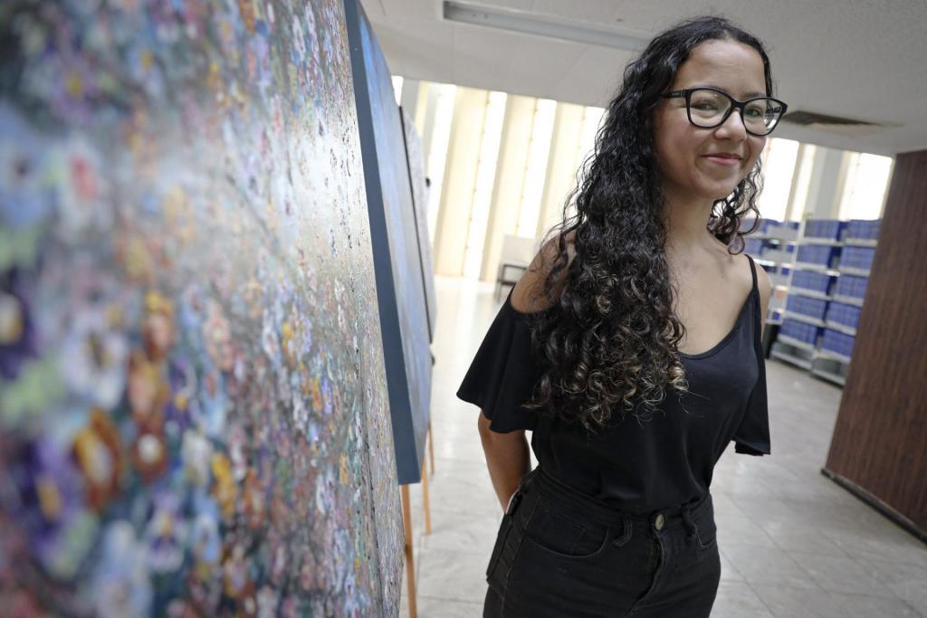 Foto colorida horizontal de Francine, uma moça com estatura mediana, cabelos cacheados e óculos. Ela veste uma roupa escura. Do lado esquerdo um painel colorido