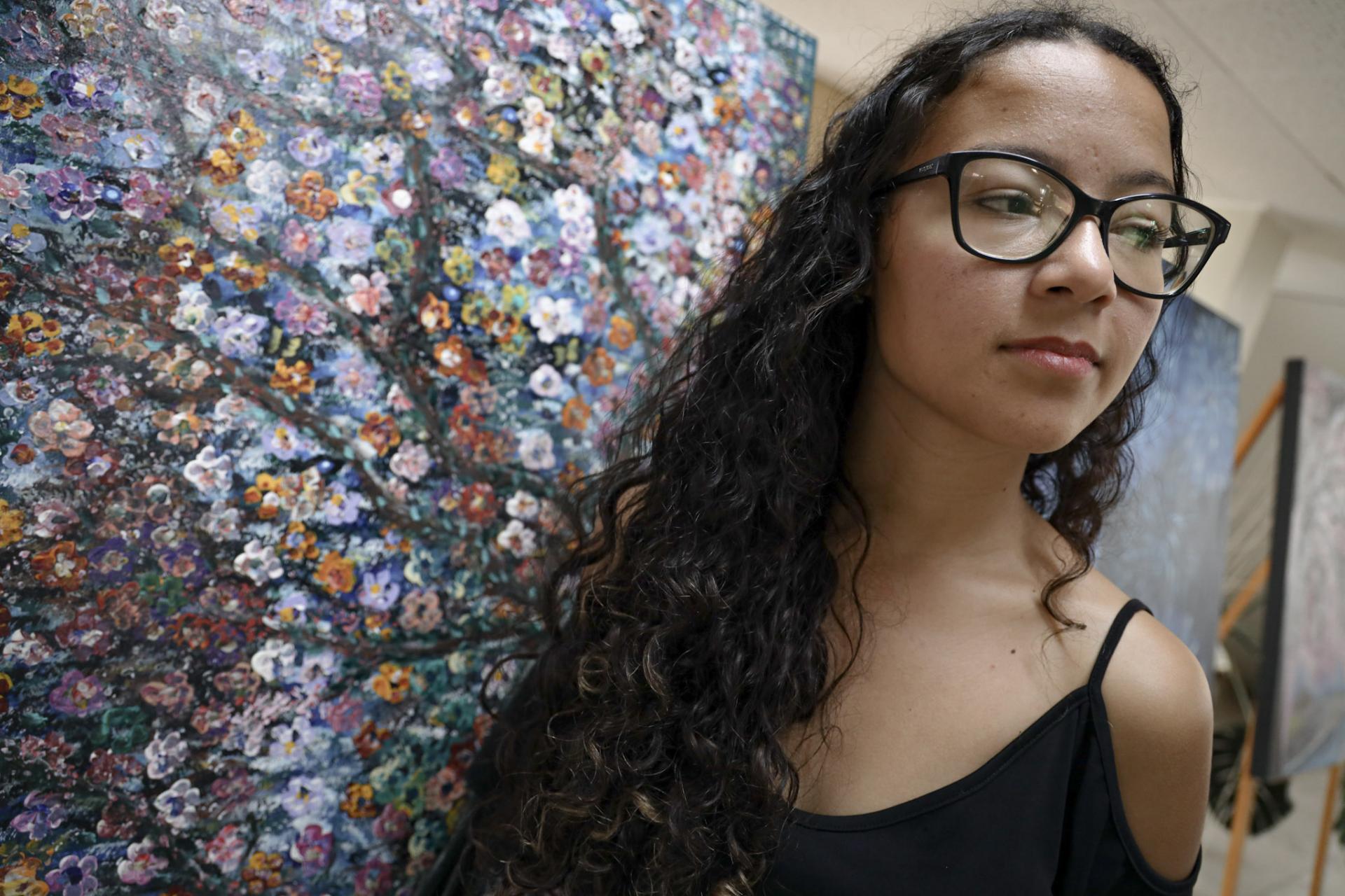 Foto colorida horizontal de Francine. A imagem traz o rosto da estudante, que está posicionada de forma inclinada à frente de um painel colorido da Biblioteca. No painel, uma árvore colorida