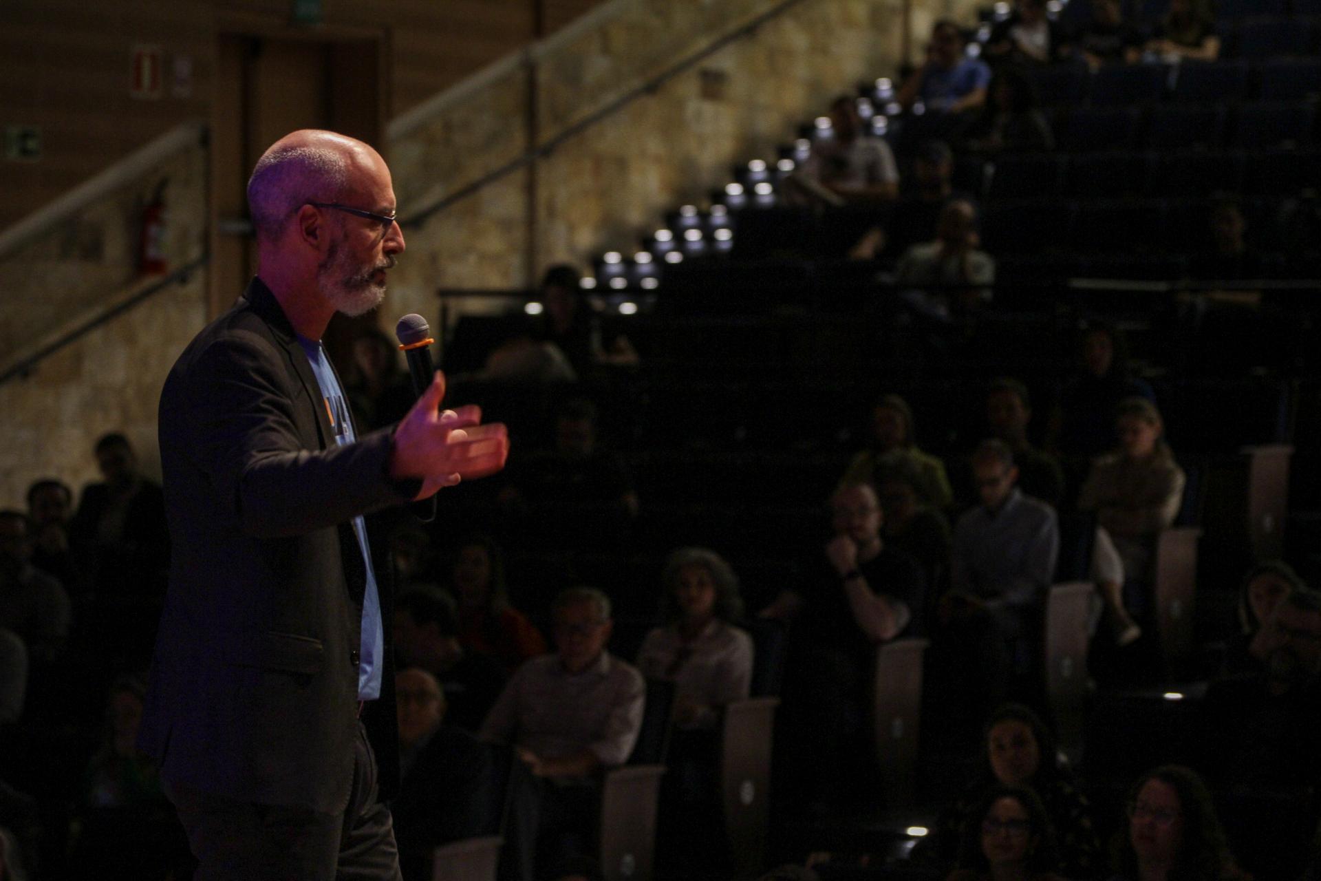 Foto colorida horizontal do reitor posicionado de lado com a mão estendida. À frente dele, o auditório lotado ao longo da plateia