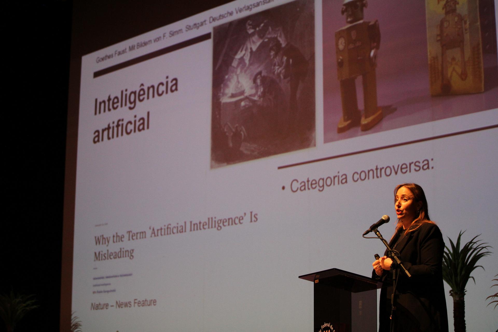 Foto horizontal. No canto direito inferior da imagem, aparece uma mulher branca de cabelos castanhos longos, vestida de preto, que está falando ao microfone. Ao fundo, aparece um slide sobre inteligência artificial, tema de sua palestra.