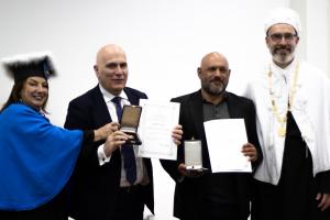 Quatro pessoas posam sorrindo durante uma cerimônia formal de entrega de honrarias. Todas estão em pé diante de um fundo branco. À esquerda, uma mulher veste uma beca azul vibrante com detalhes em preto e um chapéu cerimonial preto com uma borda branca de plumas. Ela sorri amplamente enquanto segura, ao lado do homem ao seu lado, uma caixa de medalha aberta. Ao lado dela, o segundo personagem é um homem calvo, vestido com terno escuro e gravata azul. Ele segura com as duas mãos um certificado e uma caixa de medalha aberta, mostrando o conteúdo dourado. Ele sorri discretamente, posando para a foto. À direita dele está outro homem, de barba e cabeça raspada, usando um terno preto sem gravata. Ele segura um documento de tamanho A4 e uma caixa de medalha fechada. Ele olha para a câmera com expressão tranquila.