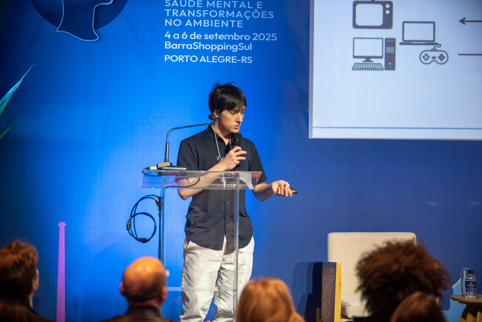 Foto colorida horizontal do estudante Ricardo em pé com microfone na mão direita, próximo à boca, e controle na mão esquerda, usado para comandar a apresentação visual projetada no telão. Atrás dele, um painel azul e um parte da apresentação com desenhos de telas. À frente dele, algumas pessoas