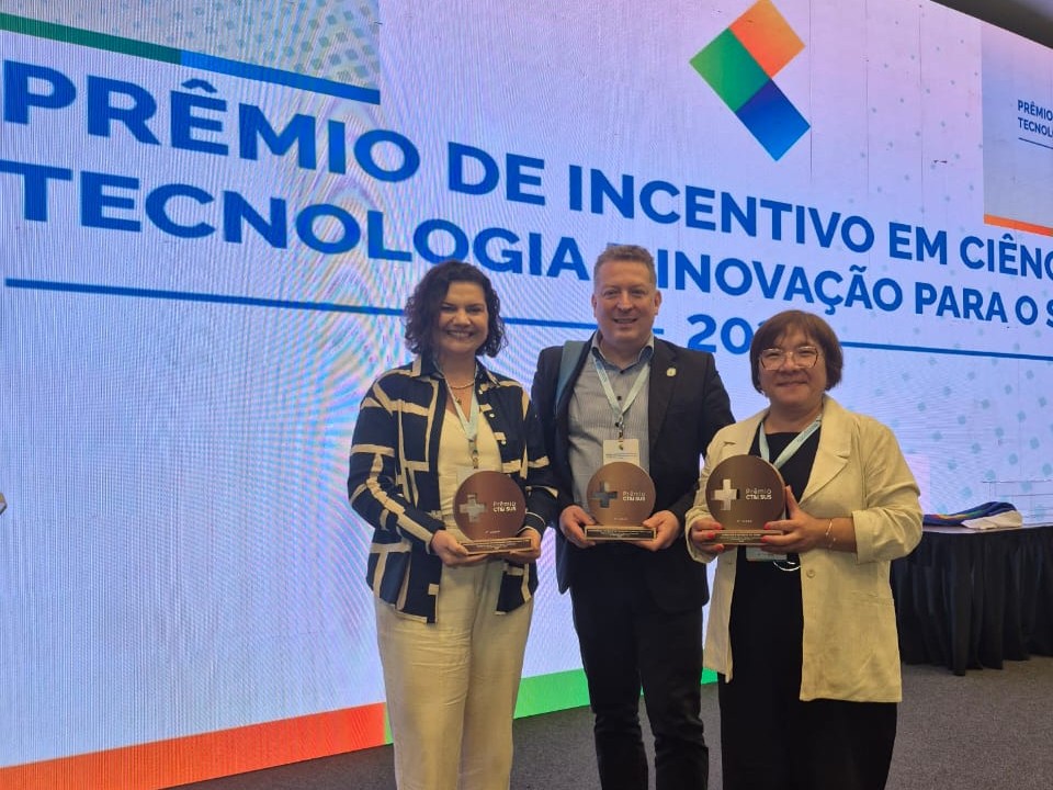 Foto vertical e colorida de duas mulheres e um homem em frente a um painel de apresentação
