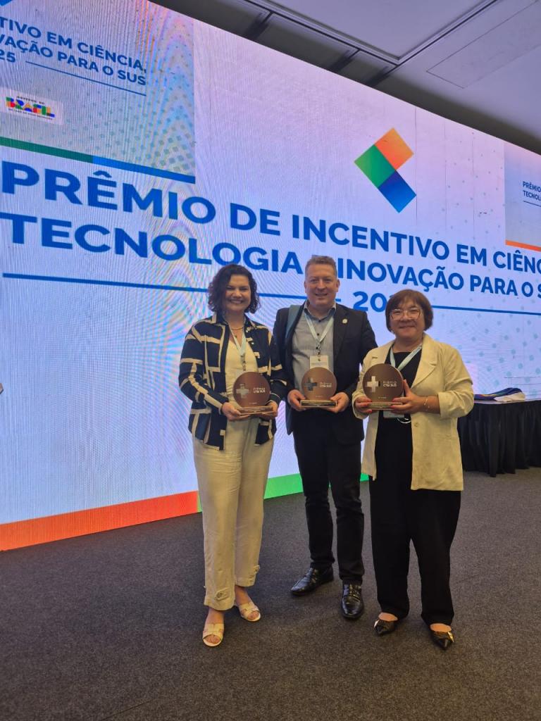 Foto vertical e colorida de duas mulheres e um homem em frente a um painel de apresentação