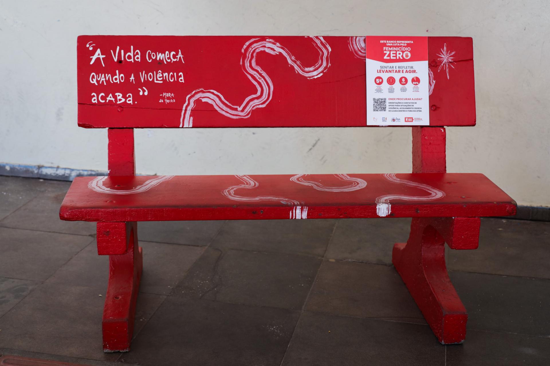 A fotografia é colorida e horizontal. A figura principal é um banco de cor vermelha com desenhos e escritas brancas. Ao lado esquerda está escrito "a vida começa quando a violência acaba", frase de Maria da Penha. Ao lado direito há uma placa com informações sobre violência contra mulheres