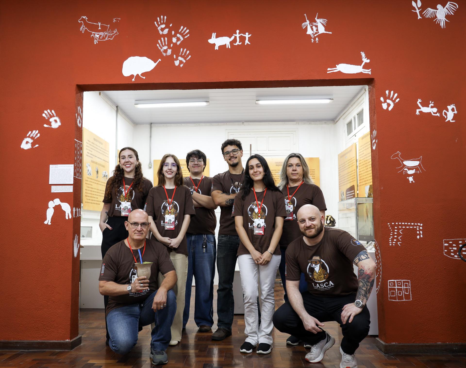 A imagem mostra um grupo de pessoas posando para uma foto. Ao redor deles, há uma parede laranja, com diversas pinturas brancas. São oito pessoas, quatro homens e quatro mulheres. Seis estão em pé, e dois homens estão ajoelhados, um de cada lado em frente ao grupo em pé. Todos usam camisetas marrons e crachás.