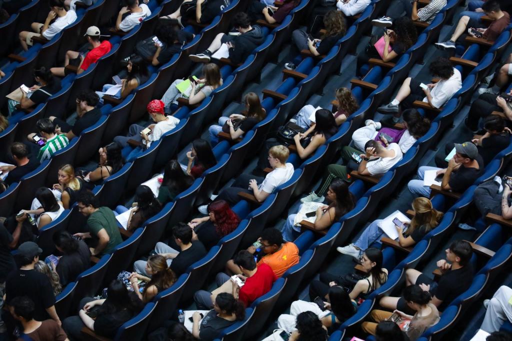 Foto colorida horizontal. A foto apresenta uma vista superior, inclinada e parcial da plateia do Centro de Convenções. A imagem mostra praticamente todas as poltronas ocupadas por pessoas que aguardam a chamada oral.