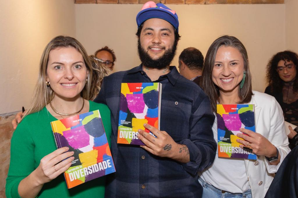 A foto mostra três pessoas: duas mulheres (à esquerda e à direita) e um homem (ao centro). Casa um deles segura um exemplar do livro lançado.