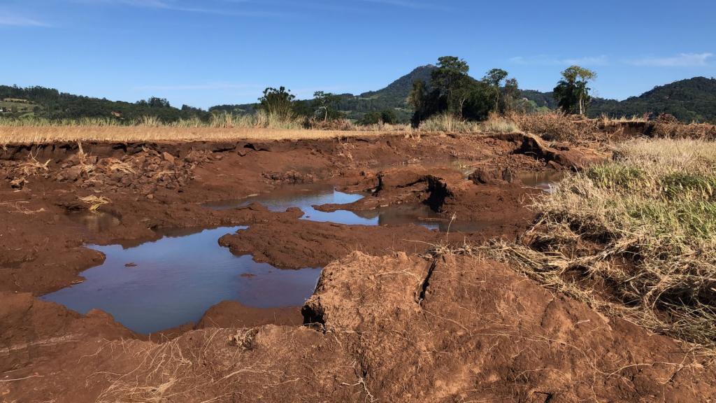 Foto horizontal colorida mostra uma área de plantação de arroz destruída pela força da água após enchentes. O solo aparece profundamente erodido, com grandes crateras preenchidas por água barrenta no centro da imagem. Ao fundo, é possível ver vegetação, árvores e montanhas sob um céu azul.