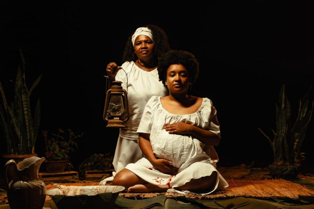 Foto colorida horizal de duas mulheres negras vestidas de branco. A primeira est谩 gr谩vida, sentada e segura a barriga. A segunda est谩 atr谩s da mulher gr谩vida e segura um lampi茫o. O vestido da mulher gr谩vida apresenta manchas vermelhos.