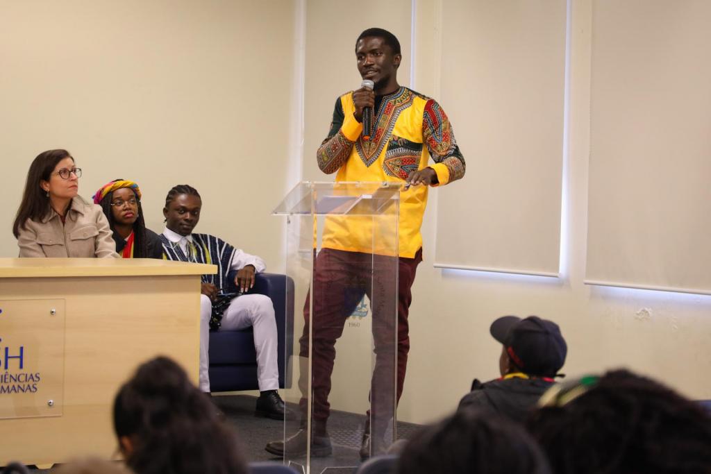 Foto colorida na horizontal. No evento de celebração ao Dia de África, Jamiro Paulo Sanca está em pé atrás de um púlpito transparente, segurando um microfone enquanto faz uma fala ao público. Ele veste uma camisa amarela com estampas africanas coloridas nas mangas e na região do peito, além de calça marrom. Ao fundo, sentados em um palco, estão uma mulher e dois estudantes observando sua fala. Aparecem parcialmente os participantes do evento voltados para o palestrante. O ambiente é um auditório com paredes claras e cortinas nas janelas.