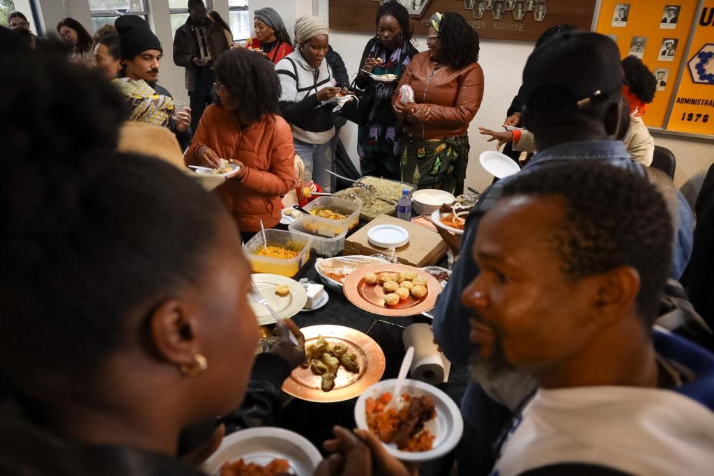 A imagem mostra um momento de confraternização em um evento cultural dedicado à gastronomia africana. Ao redor de uma mesa repleta de pratos típicos, diversas pessoas conversam, servem-se e compartilham a refeição em um ambiente de integração e troca cultural. Sobre a mesa, é possível observar diferentes preparações tradicionais, enquanto os participantes circulam pelo espaço, degustando os alimentos e interagindo entre si. A cena destaca a diversidade, a convivência e a valorização das tradições culinárias africanas por meio da experiência coletiva da comida.