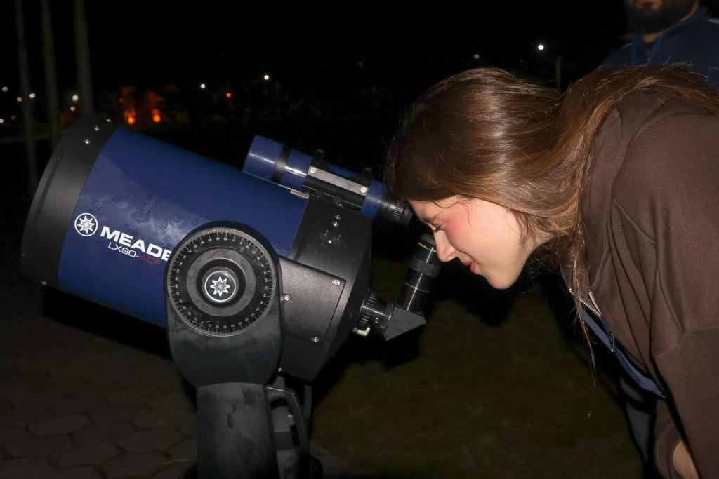 Uma moça observa o céu à noite por meio de um telescópio. Ela é branca e tem cabelo castanho, longo e liso. Está vestida com um casaco marrom.