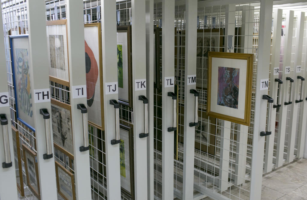 Fotografia horizontal e colorida de uma estrutura de madeira e metal em formato de gavetas verticais. São 12 gavetas, com puxador de metal cinza e preto, com letras do alfabeto coladas em cada uma. Uma das gavetas, à direita da imagem, está puxada para fora. Nela, um quadro de moldura escura está preso em uma estrutura de metal, no interior da gaveta, em branco.