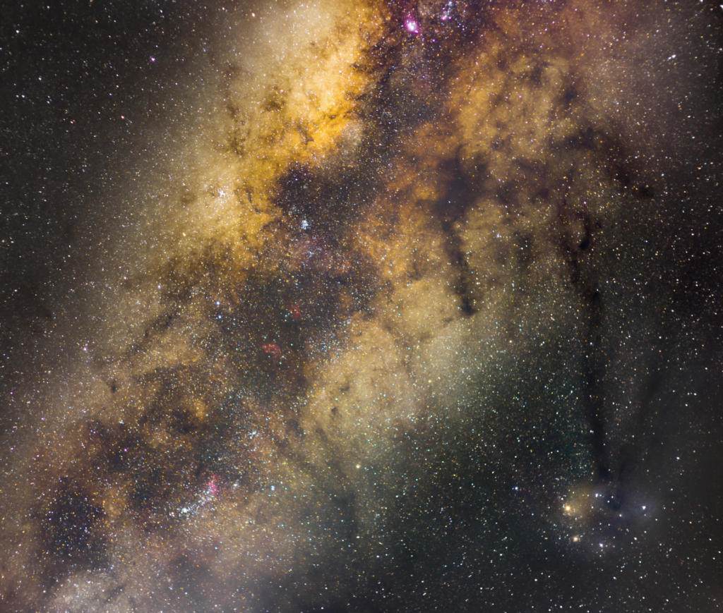 Fotografia horizontal e colorida da Via Láctea em tons de roxo, azul escuro, preto e amarelo esverdeado. Há pontos de luz espalhados pela fotografia..