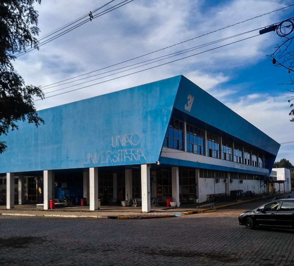 Descrição de Imagem: Fotografia horizontal e colorida da diagonal de um prédio de dois andares na cor azul. Embaixo, no primeiro andar, nove pilares de sustentação na cor branca; a fachada é azul e, na parte inferior direita da fachada, está escrito "UNIÃO UNIVERSITÁRIA”, em caixa alta, letras grandes e na cor branca. No lado direito do prédio, há janelas espelhadas e “31” na ponta, em cor branca. Na parte superior, o céu em azul com nuvens brancas. No chão, pedras em paralelepípedos na cor cinza. Um carro preto em movimento se direciona para a rua localizada ao lado direito do prédio.