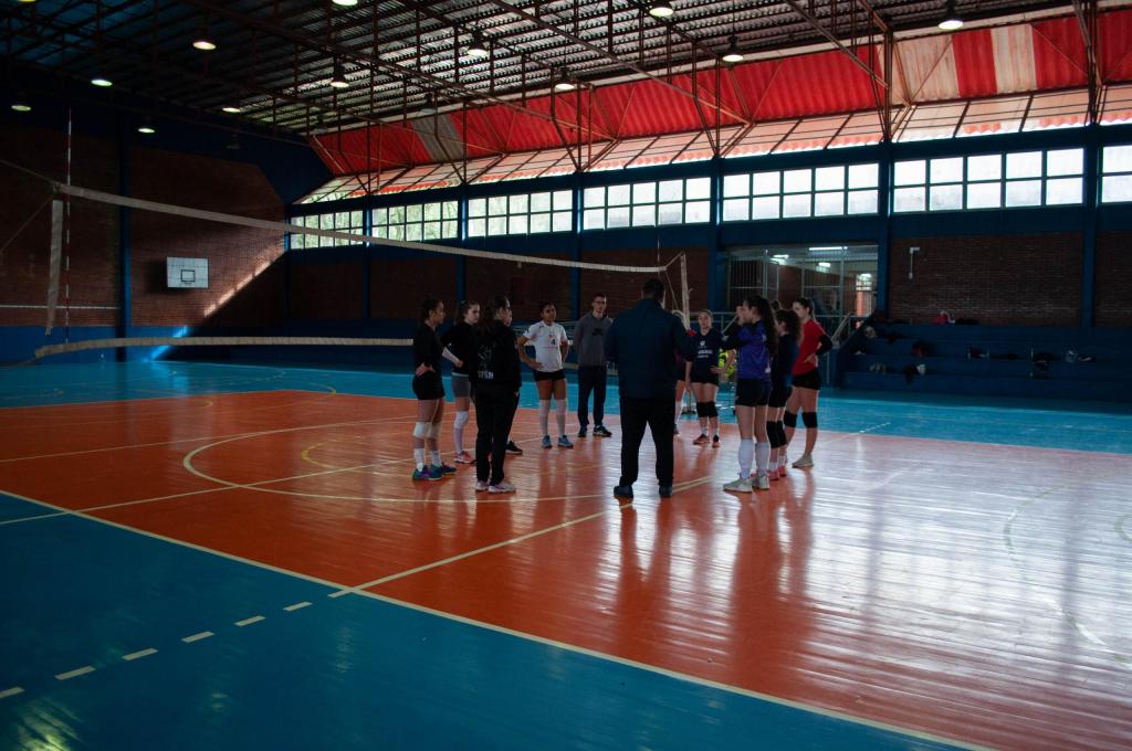 Foto em sentido horizontal, colorida, com um círculo formado pelas atletas e comissão técnica em destaque, dentro de uma quadra esportiva. Ao fundo, está a quadra de vôlei na cor laranja e as extremidades na cor azul, a rede de vôlei transparente com bordas brancas e dez atletas, todas mulheres que estão em pé, assim como dois homens, que também estão em pé. 