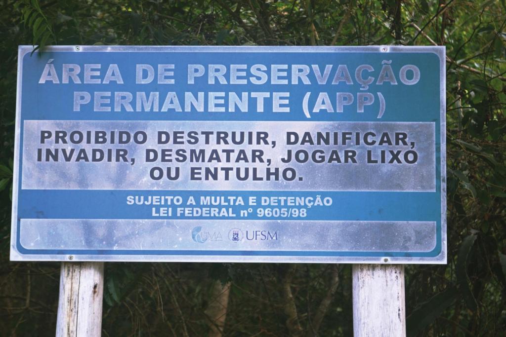 Fotografia horizontal, colorida, em primeiro plano e ângulo fechado, de uma placa que delimita uma Área de Preservação Permanente. A placa tem tem detalhes em azul e prata fixada em um suporte de madeira. Está com limo e suja, ao fundo, há árvores que fecham o ambiente ao redor. A iluminação é natural.