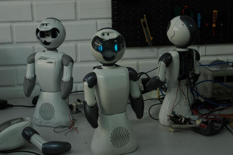 Fotografia horizontal, colorida em plano médio de três robôs humanoides pequenos sobre uma bancada branca. Eles estão em pé, um ao lado do outro, e têm corpos brancos, cabeça arredondada e dois braços, com articulações em cinza-escuro. Eles não têm pernas, apenas uma base larga de sustentação, em que há uma abertura para alto-falante formada por linhas sinuosas. Também não têm mãos, mas um alongamento do braço, em cinza escuro. O robô no lado esquerdo da imagem está danificado: não tem o visor frontal montado, é possível ver os componentes internos e há marcas de desgaste no corpo. O robô do centro está ligado, e no visor preto tem olhos formados por dois pontos de luz azul. O robô do lado direito da imagem está de costas,aberto, de onde saem fios e componentes eletrônicos. Sobre toda a bancada estão cabos pretos, vermelhos e outros componentes eletrônicos. Ao fundo, uma parede de tijolos brancos e uma prateleira com ferramentas.