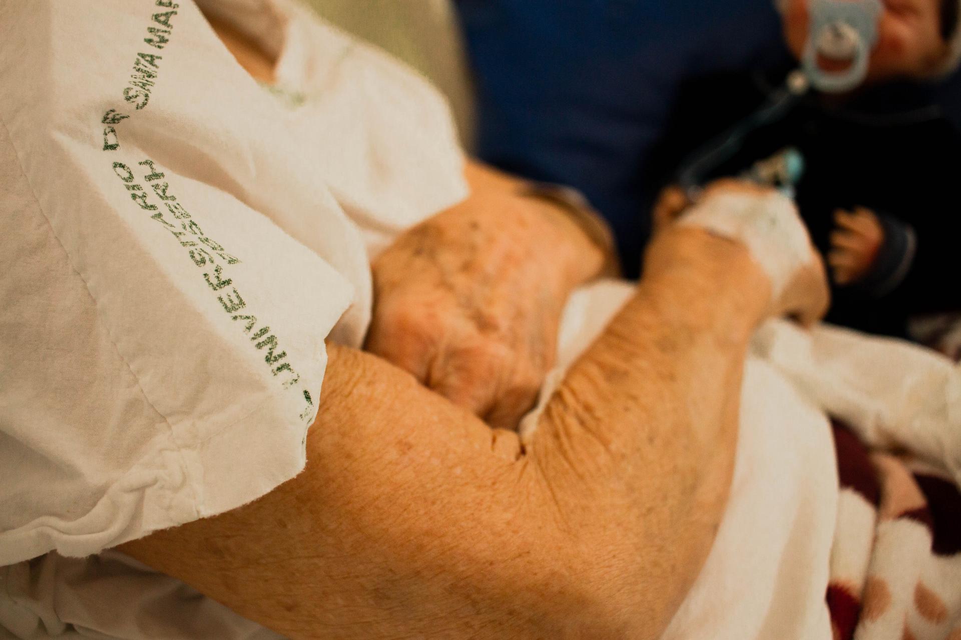 Fotografia horizontal, colorida e em plano detalhe da pele de Neite, que enfoca as marcas da idade. A pele é branca. A idosa parece estar na faixa etária dos 80 a 90 anos. Na mão esquerda há um acesso venoso, fixado por um pedaço de fita. Está sentada em uma maca hospitalar e veste um avental na cor branca, com o nome “Hospital Universitário de Santa Maria” escrito na cor verde. Ela está coberta da cintura para baixo por uma manta branca com detalhes em vermelho. Ao fundo da imagem, desfocado, está um boneco sentado sobre uma almofada azul marinho. Ele tem a pele branca, veste um tip top azul marinho e está com uma chupeta azul claro na boca.