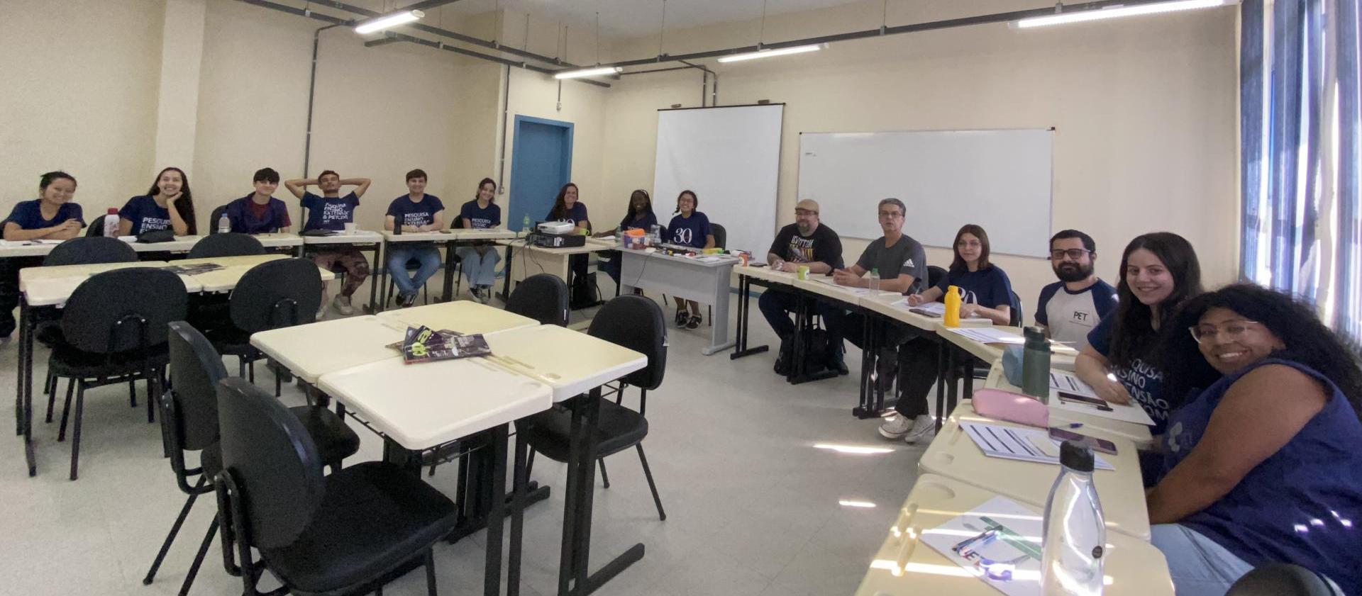 Foto de uma sala de aula, com mesas e cadeiras posicionadas em formato de meia lua. Nas cadeiras estão sentados 12 integrantes do PETCom, utilizando camisetas de cor azul-marinho com texto em branco, onde se lê "Ensino. Pesquisa. Extensão. E PETCom". Também há a presença de três professores homens e uma professora mulher. Ao fundo, um quadro branco e uma porta na cor azul.